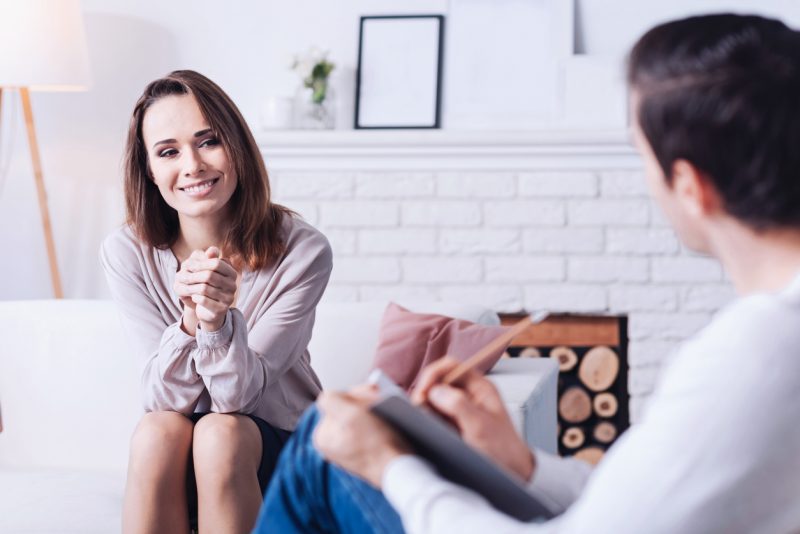 Woman seated on a couch, elbows on her knees and clasped hands, smiling and engaged in conversation with her hypnotherapist who is taking notes.