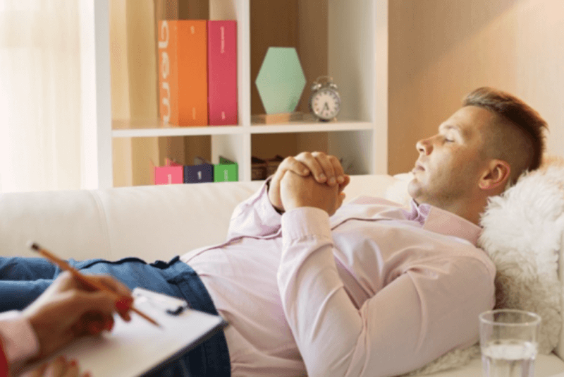 Man lying on a couch during a hypnotherapy session whilst the therapist is taking notes